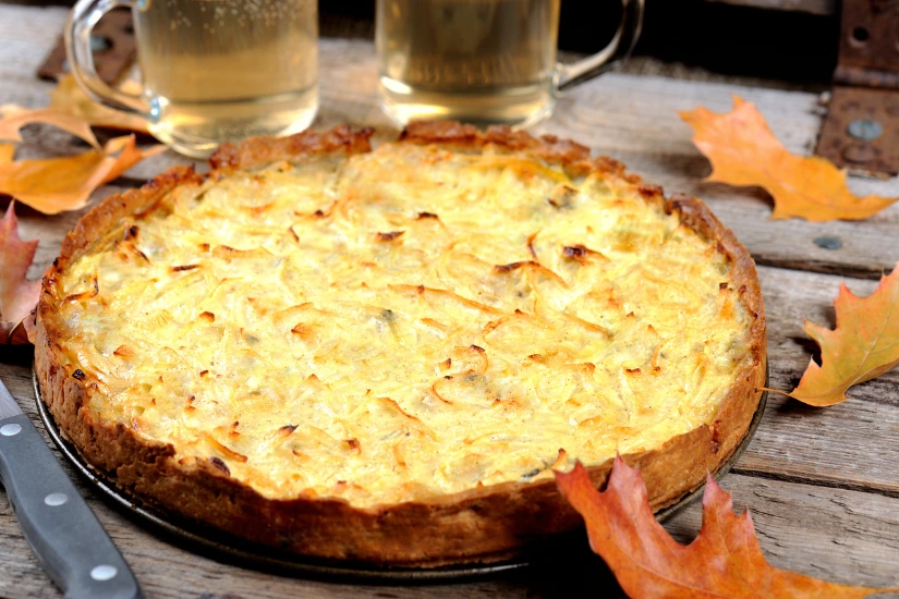 Herzhafter Zwiebelkuchen auf einem rustikalen Holztisch mit Herbstblättern und zwei Gläsern Apfelwein.